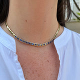 A close-up of a 14K yellow gold tennis-style choker necklace with rectangular blue sapphires, worn by a person wearing a white collared shirt.