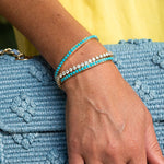 Two gold tennis-style bracelets worn on a woman's wrist, one with bright turquoise blue beads and the other with clear diamonds, resting on a textured light blue bag.