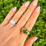 A close-up of a hand wearing two rings against a blurred green foliage background. The primary product shown is an 18K gold signet-style ring with a green oval tourmaline gemstone, with another silver ring also visible on the opposite finger.