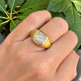 A gold signet-style ring with a rectangular emerald-cut clear diamond, shown worn on a woman's hand with green leaves in the background.