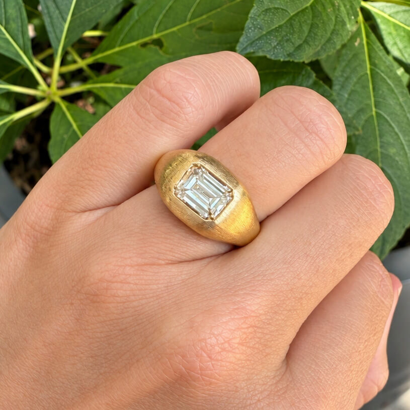 A gold signet-style ring with a rectangular emerald-cut clear diamond, shown worn on a woman's hand with green leaves in the background.