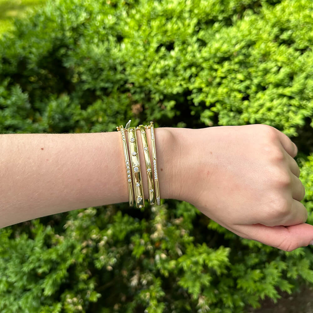 Hand wearing multiple gold bracelets with a green bush background