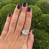 Hand wearing an 18k gold ring with a large moon quartz gemstone against a green mossy background