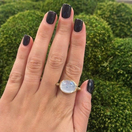 Hand wearing an 18k gold ring with a large moon quartz gemstone against a green mossy background