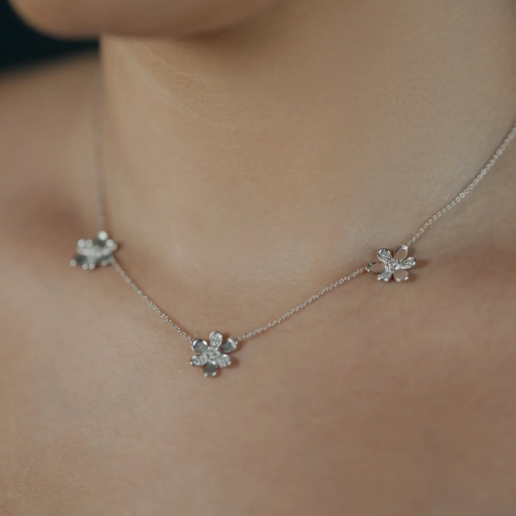 A close-up of a delicate 18k white gold chain necklace worn by a model, featuring three small, flower-like charms embellished with pave diamonds