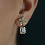 A close-up of a woman's ear wearing an 18K yellow gold flower-shaped stud earring adorned with small clear diamonds and a dangling rectangular blush colored amethyst gemstone.