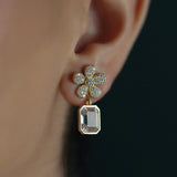 A close-up of a woman's ear wearing an 18K yellow gold flower-shaped stud earring adorned with small clear diamonds and a dangling rectangular blush colored amethyst gemstone.