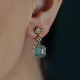 A close-up of a woman's ear wearing an 18K yellow gold drop earring with two small clear diamonds in square settings and a larger rounded pale blue-green cabochon aquamarine stone.