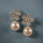 A pair of flower-shaped drop earrings with pavé-set clear stones and large round pearl-like drops, photographed on a soft blue-gray textured background.