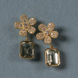 A pair of 18K yellow gold flower-shaped drop earrings with pavé-set clear diamonds and large rectangular green amethyst gemstones hanging below, displayed on a textured blue-gray background.