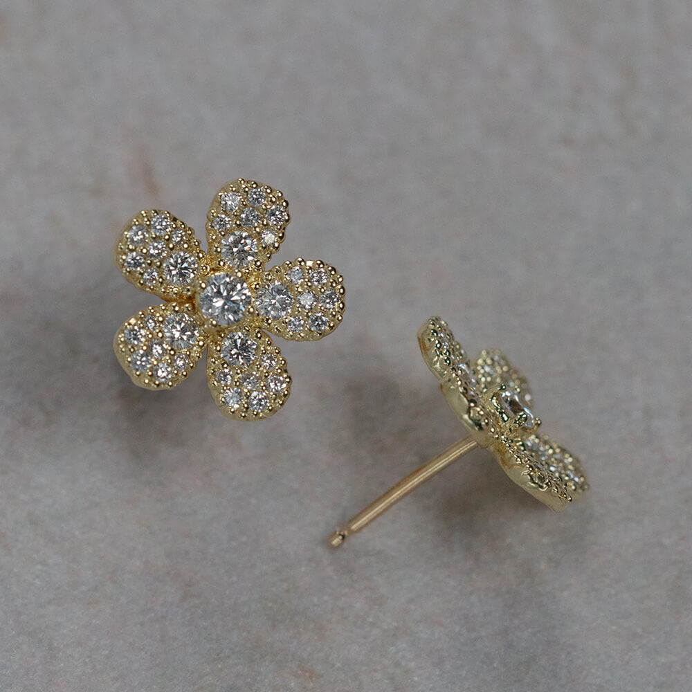 A pair of 18K yellow gold flower-shaped stud earrings paved with clear diamonds, shown on a neutral textured gray background. One earring is shown from the front, displaying the five-petal flower design, and the other is shown from the side/back, displaying the post and backing.