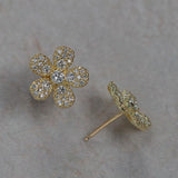 A pair of 18K yellow gold flower-shaped stud earrings paved with clear diamonds, shown on a neutral textured gray background. One earring is shown from the front, displaying the five-petal flower design, and the other is shown from the side/back, displaying the post and backing.