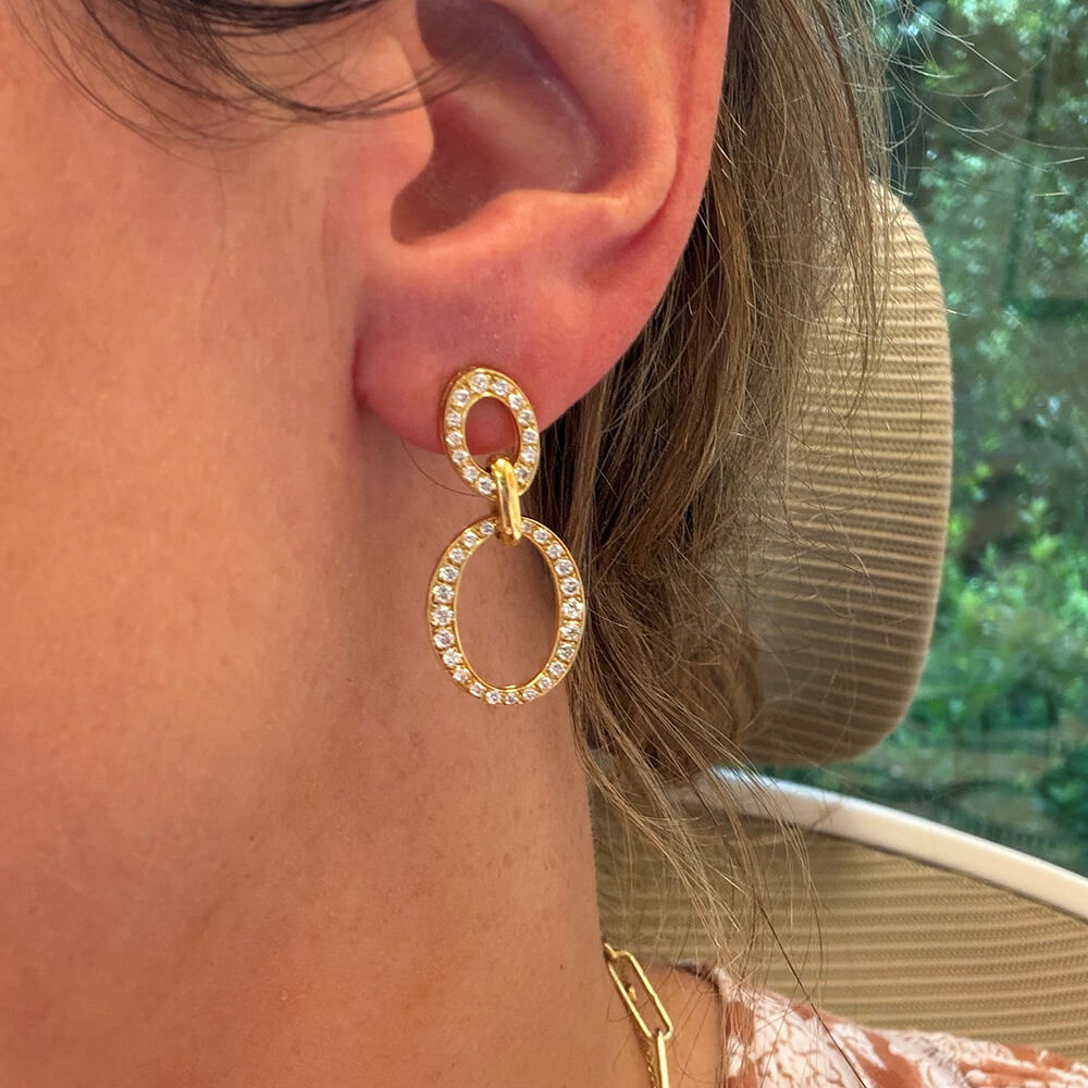 A close-up of a woman's ear wearing an 18K gold drop earring adorned with clear diamonds, 