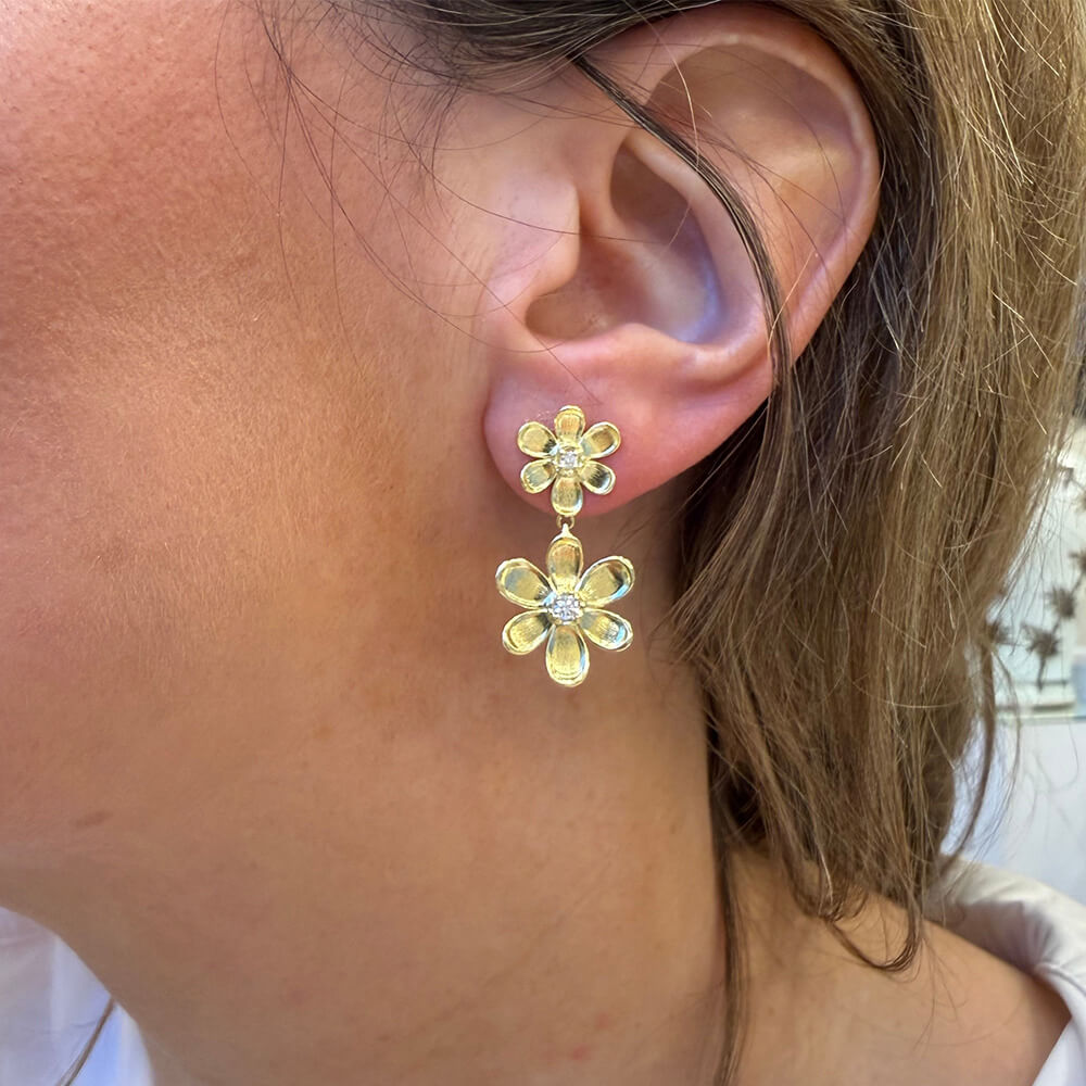 A close-up of a woman's ear wearing an 18K yellow gold double flower drop earrings with diamond centers