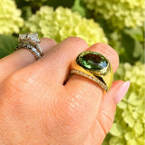 A close-up of a hand wearing an 18K gold signet-style ring with a large oval green tourmaline gemstone, alongside another silver ring, against a soft-focus background of light green hydrangeas.