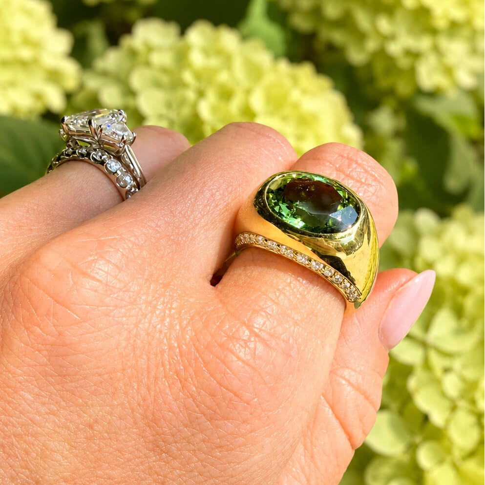 A close-up of a hand wearing an 18K gold signet-style ring with a large oval green tourmaline gemstone, alongside another silver ring, against a soft-focus background of light green hydrangeas.