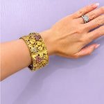 A close-up of a woman's hand and wrist wearing an ornate wide 18K yellow gold cuff bracelet decorated with vine and grape motifs, accented with rubies and diamonds