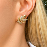 A close-up of a woman's ear wearing an 18K gold leaf-shaped stud earring adorned with small clear diamonds.