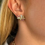 A close-up of a woman's ear wearing an 18k gold leaf-shaped stud earring adorned with clear pavé diamonds and pink sapphire accents. 