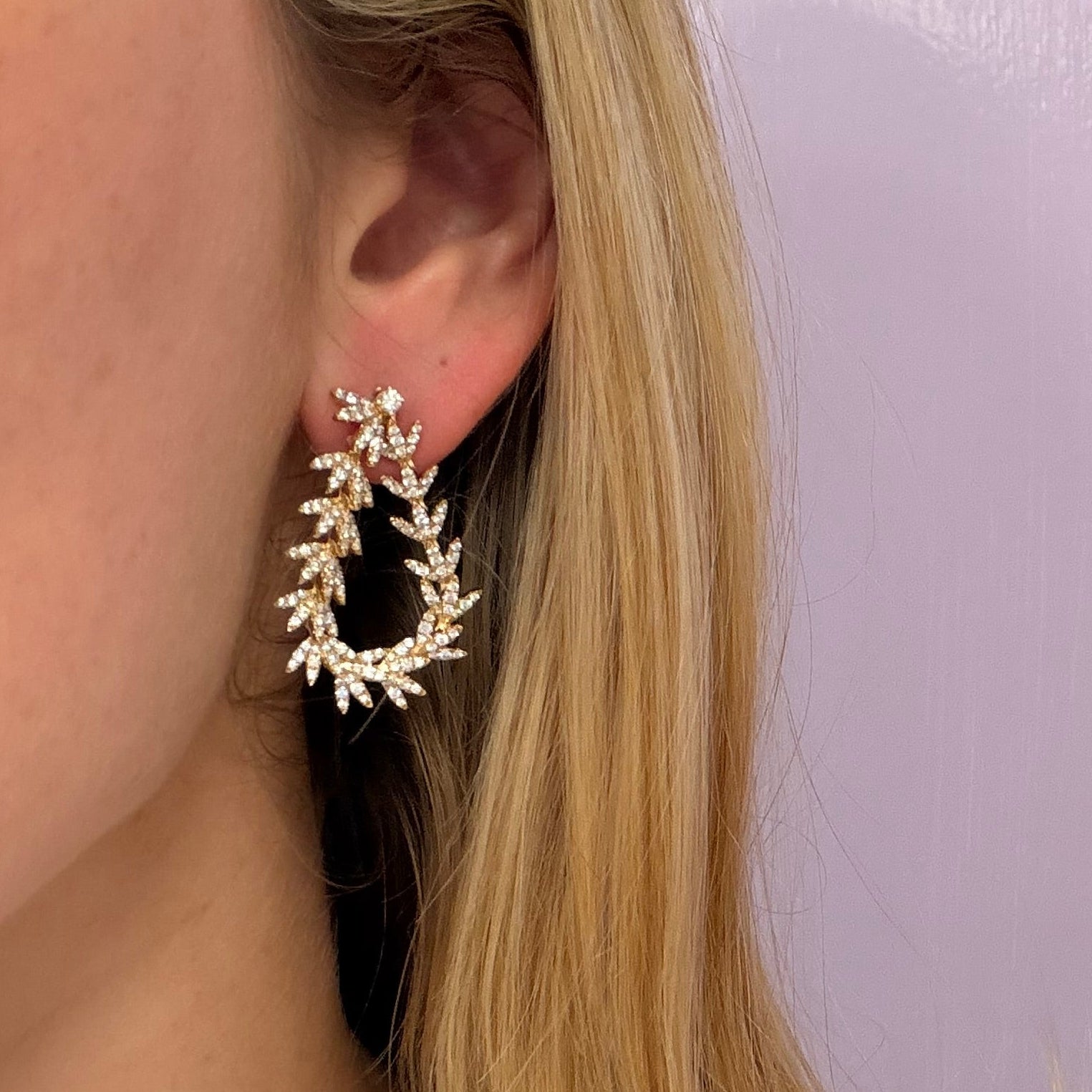 Close-up of a woman wearing an 18k gold hoop dangle diamond earring against a light purple background 