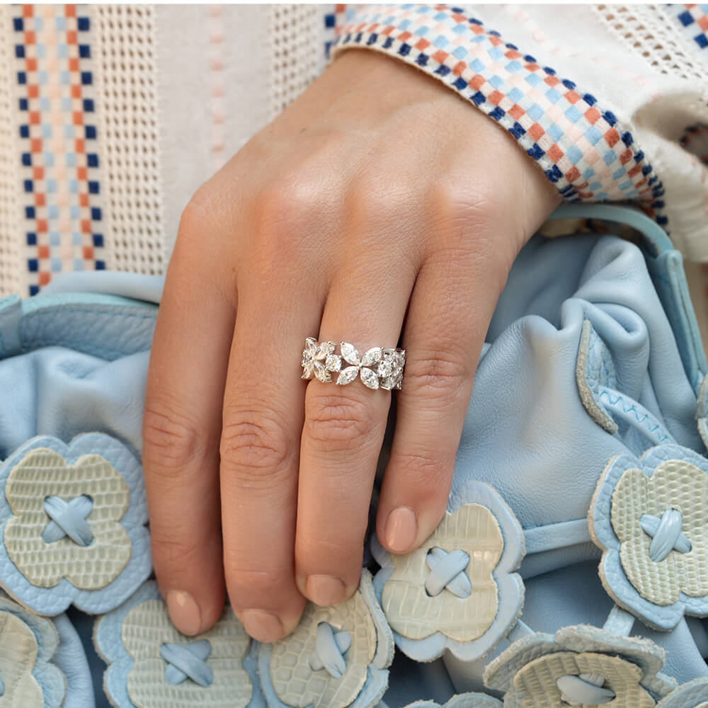 A close-up of a woman's hand wearing an 18K white gold ring with a delicate floral leaf pattern of marquise diamonds, resting on a light blue textured fabric bag.