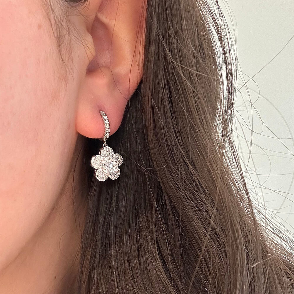 A close-up of a woman's ear wearing an 18K white gold huggie hoop earring with a dangling flower-shaped diamond cluster.