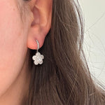 A close-up of a woman's ear wearing an 18K white gold huggie hoop earring with a dangling flower-shaped diamond cluster.