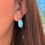 A close-up of a woman's ear wearing a drop earring with a small diamond stud and a large oval light blue turquoise dangle.