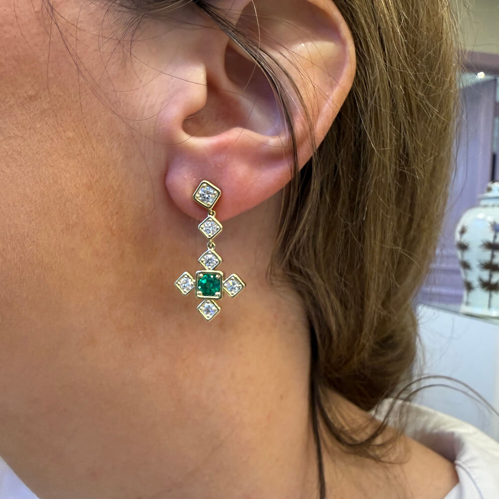 A close-up of a woman's ear wearing an 18K gold dangle earring with a central green emerald and several diamonds. 