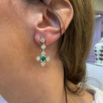 A close-up of a woman's ear wearing an 18K gold dangle earring with a central green emerald and several diamonds. 