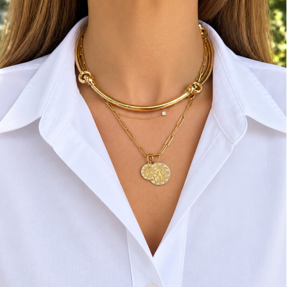 A close-up of a model wearing a layered 18K yellow gold necklace set, featuring a thick bangle-style top layer, a delicate chain with a small diamond bead, and a lower chain with a textured gold coin pendant. The model is wearing a white collared shirt.