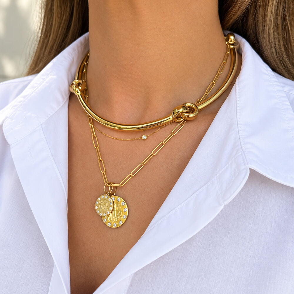 A close-up of a woman wearing a layered 18K yellow gold necklace set with a thick curved chain, a delicate chain with a small diamond bead, and a round gold pendant with a white floral pattern.