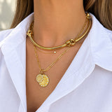 A close-up of a woman wearing a layered 18K yellow gold necklace set with a thick curved chain, a delicate chain with a small diamond bead, and a round gold pendant with a white floral pattern.