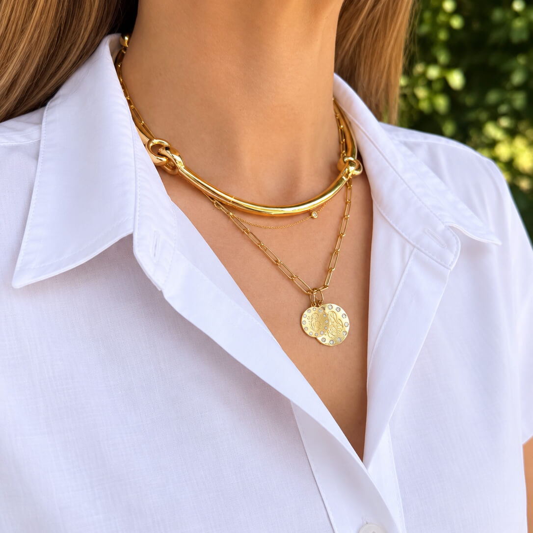 A close-up of a woman wearing layered 18K yellow gold necklaces, including a thick curved bar necklace, a delicate chain necklace with a textured oval pendant, and a small diamond, paired with a white collared shirt.