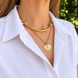 A close-up of a woman wearing layered 18K yellow gold necklaces, including a thick curved bar necklace, a delicate chain necklace with a textured oval pendant, and a small diamond, paired with a white collared shirt.