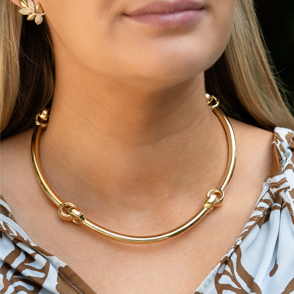 A close-up of a woman wearing a polished gold hinged collar necklace. The necklace features four interlocking circular rings and is shown worn with a brown and white patterned top.