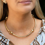 A close-up of a woman wearing a polished gold hinged collar necklace. The necklace features four interlocking circular rings and is shown worn with a brown and white patterned top.