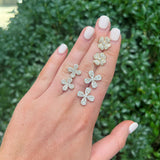 A hand with white manicured nails displays flower-shaped stud earrings set with clear diamonds, against a blurred background of green bushes.