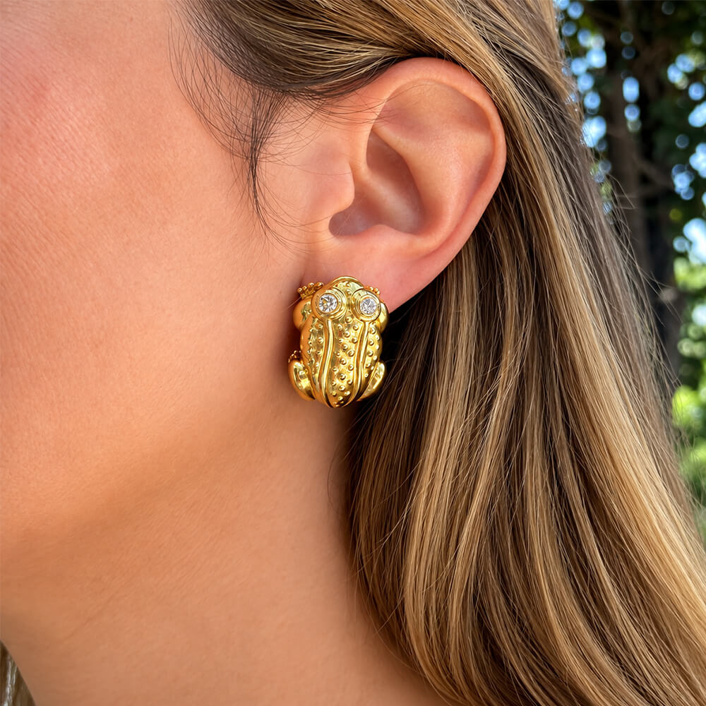 A close-up of a woman's ear wearing an estate 18k yellow gold frog-shaped earring with two small clear diamonds for the eyes, set against her hair and a blurred outdoor background.