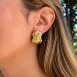 A close-up of a woman's ear wearing an estate 18k yellow gold frog-shaped earring with two small clear diamonds for the eyes, set against her hair and a blurred outdoor background.