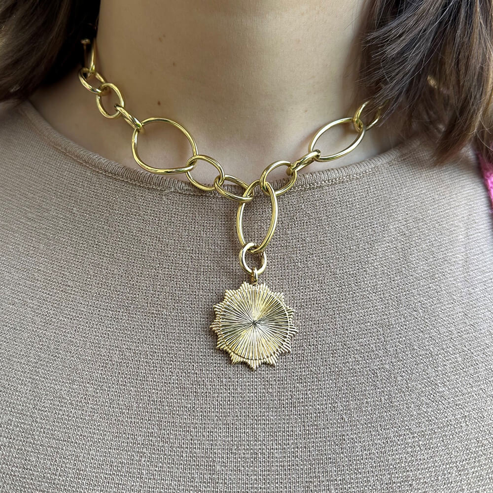 A close-up of a woman wearing a chunky gold chain necklace with a textured 18K yellow gold sunburst pendant, paired with a beige knit top.