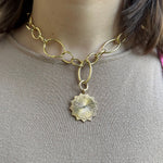 A close-up of a woman wearing a chunky gold chain necklace with a textured 18K yellow gold sunburst pendant, paired with a beige knit top.