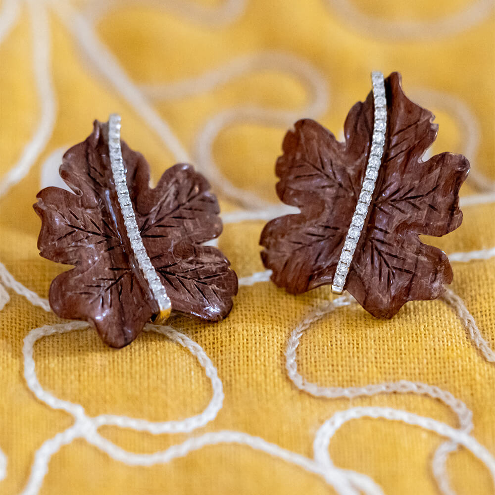 A pair of brown leaf-shaped earrings with a vertical line of small clear diamonds, displayed on a yellow embroidered fabric background.