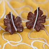 A pair of brown leaf-shaped earrings with a vertical line of small clear diamonds, displayed on a yellow embroidered fabric background.