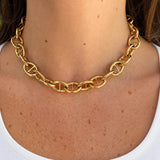 A close-up of a woman's neck and upper chest wearing a chunky 18k yellow gold chain necklace with interlocking oval mariner-style links over a white tank top.