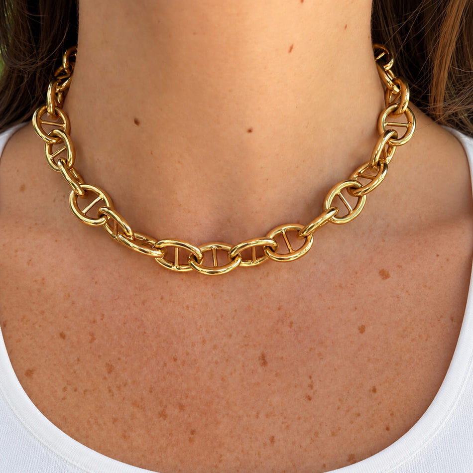A close-up of a woman's neck and upper chest wearing a chunky 18k yellow gold chain necklace with interlocking oval mariner-style links over a white tank top.