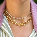 A close-up of a woman's neck wearing two layered necklaces: a thick 18k yellow gold round mariner chain and a delicate gold chain with small clear diamonds, styled with a pink textured cardigan and a green and white striped shirt.