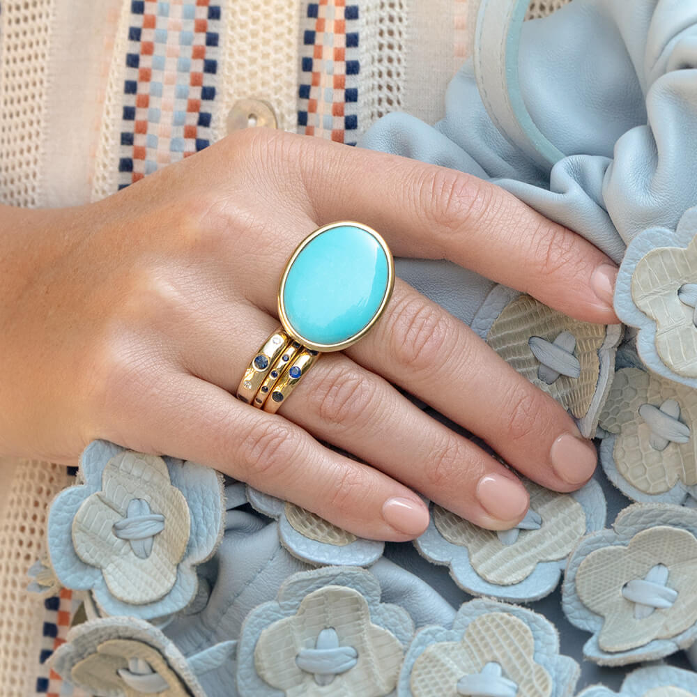 A close-up of a hand wearing a gold ring with a large oval turquoise cabochon, layered with three slim 18k yellow gold and sapphire stackable band rings, resting on a light blue textured fabric.