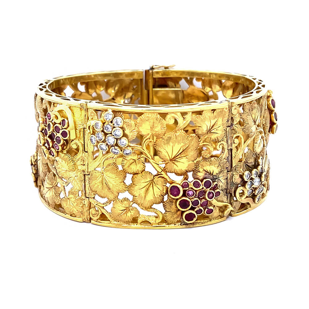 An ornate 18K yellow gold cuff bracelet featuring detailed vine and grape motifs, accented with clear diamonds and deep red rubies, shown against a plain white background.