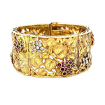 An ornate 18K yellow gold cuff bracelet featuring detailed vine and grape motifs, accented with clear diamonds and deep red rubies, shown against a plain white background.